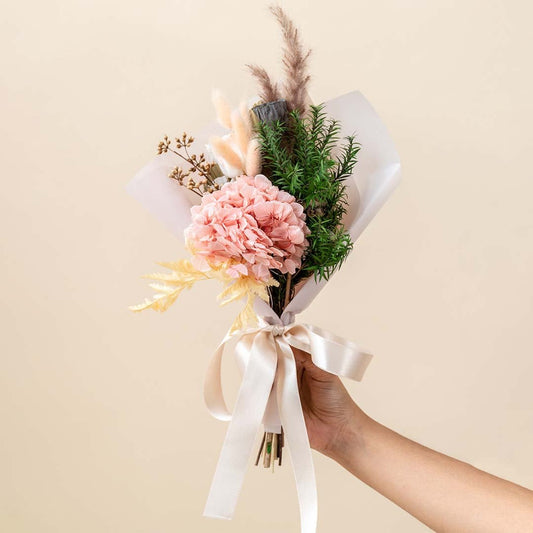 Dried flower bouquet with pink hydrangea pampas grass lotus pod and green foliage tied with cream ribbon is a preserved floral arrangement.