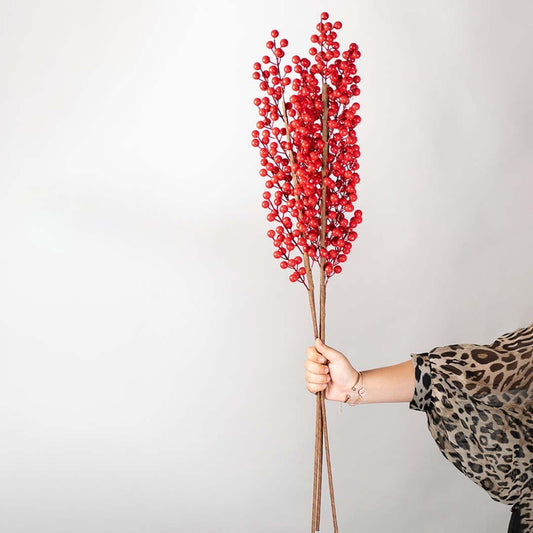 Hand holding a bundle of vibrant red artificial berry branch stems for festive holiday tabletop vase on white backdrop.