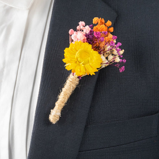 Closeup view of groom boutonniere with bright yellow artificial flower and colorful dried blooms wrapped in jute on suit.