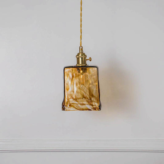 Square amber glass pendant light with gold fixture and hanging cord placed against neutral wall background enhancing modern artistic lighting decor styling.