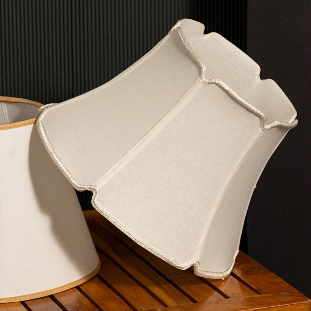A soft beige fabric lampshade with a scalloped edge design displayed on a wooden surface with a black background creating a warm side view look.