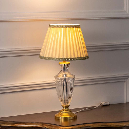 Crystal glass table lamp with gold base and beige pleated shade turned on against white background creating a warm inviting glow for bedroom decor.