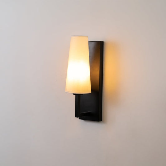 Black metal wall light with tall white fabric shade glowing softly on a clean white background ideal for modern bedroom or hallway lighting.