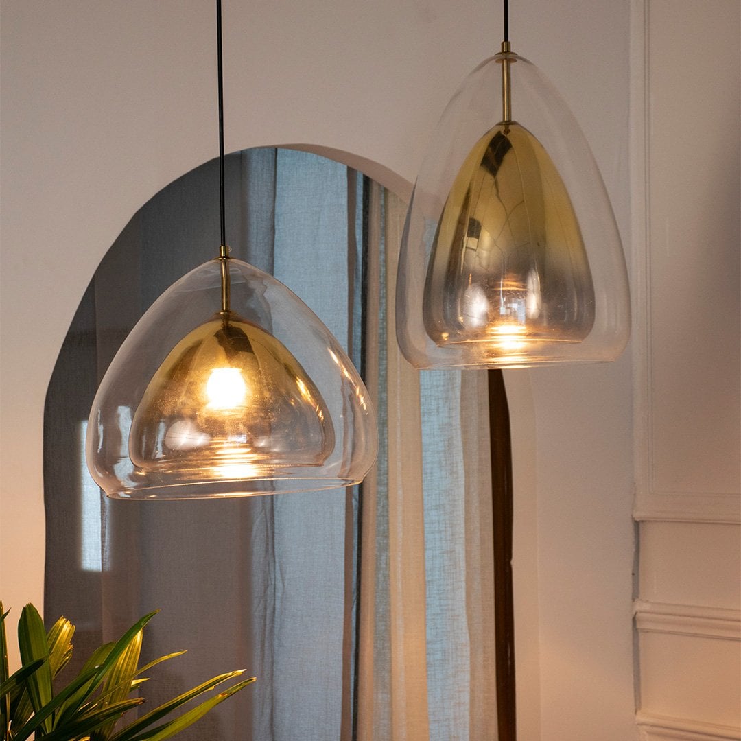 Warm pendant lights glowing in a cozy room setting with sheer curtains and indoor plants creating a soft inviting ambiance around the golden glass fixtures.