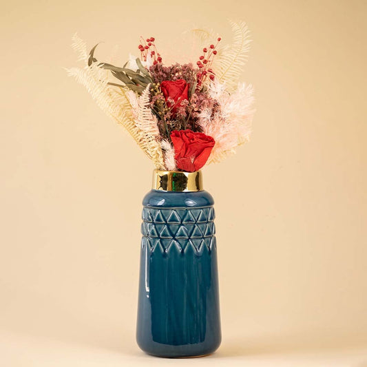 Elegant dark blue ceramic vase with a gold rim filled with red roses ferns and dried flowers placed against a warm beige background creating a rich and luxurious decorative look.