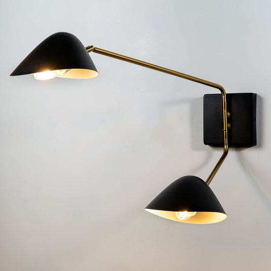 A twin head wall lamp with black shades and gold arms displayed against a white background with both lights on creating a warm glowing effect.