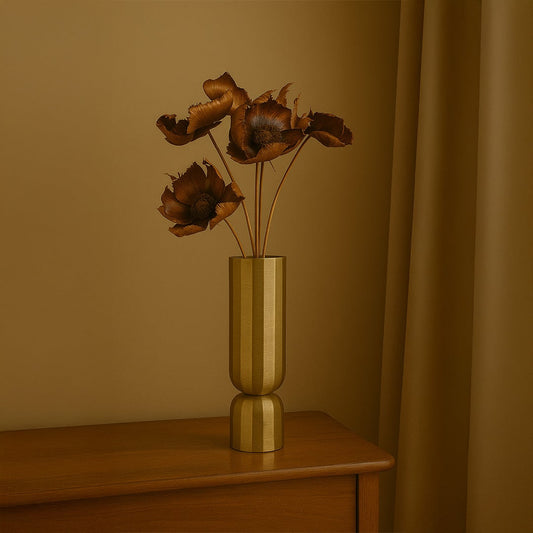 A tall gold metal vase holding dried brown flowers placed on a wooden surface against a warm beige backdrop creating elegant earthy botanical accents for sophisticated interior décor.