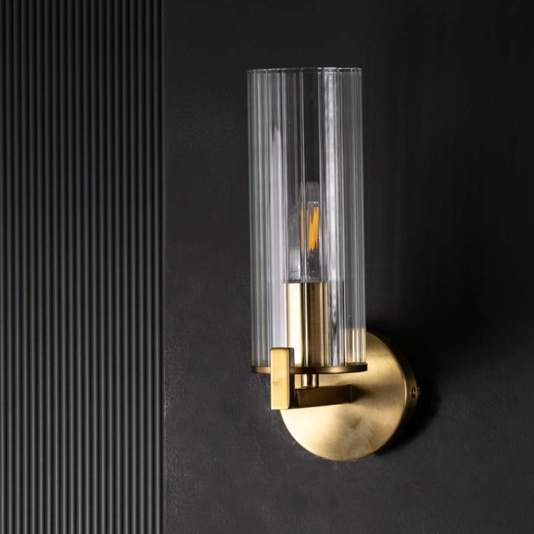 A gold wall sconce with a tall ribbed glass shade showcased on a dark background with the light off highlighting its sleek elegant form.