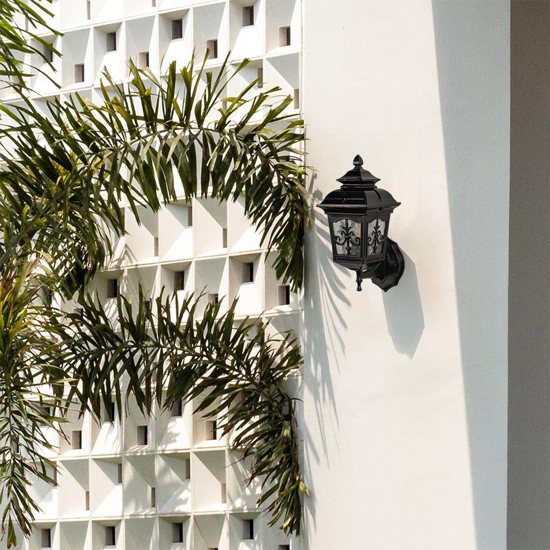 A decorative black outdoor lantern mounted on a bright exterior wall adding charm to the outdoor walkway while blending with the lush green surroundings.
