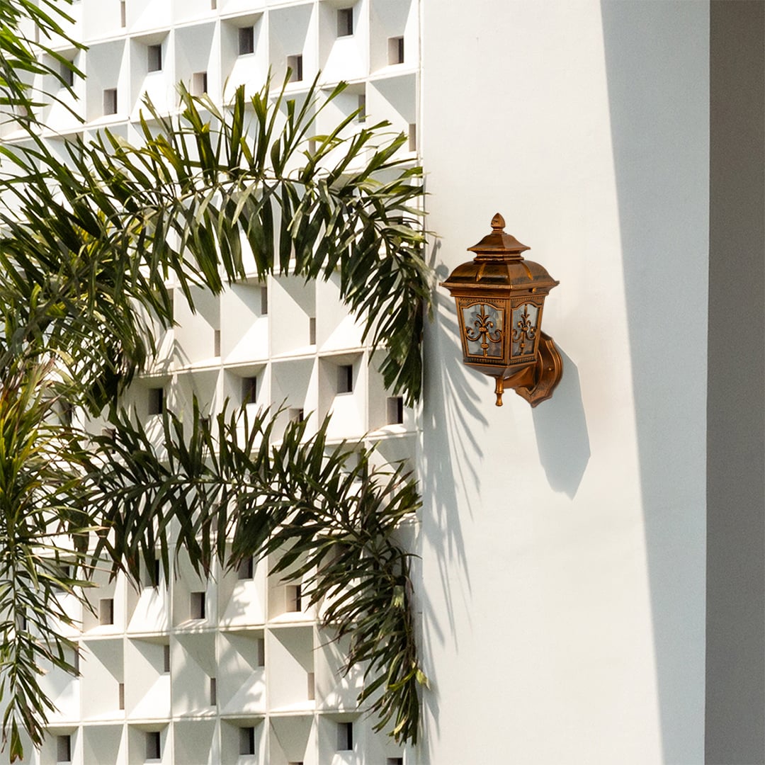 A gold outdoor wall lantern mounted on a white exterior facade adding decorative charm and subtle illumination to a bright open outdoor space in natural daylight.