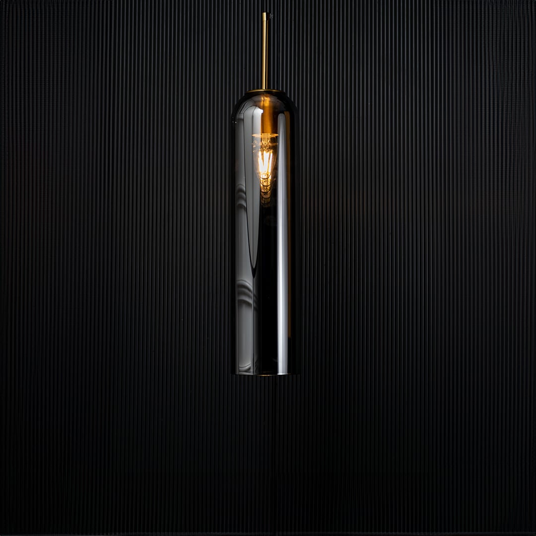A modern silver cylindrical pendant light against a deep black background with the light turned on casting a warm glow and highlighting its reflective finish.