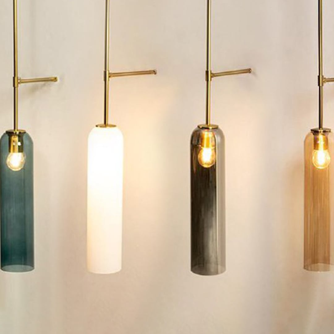 Close up of four cylindrical pendant lights with long glass shades in blue white grey and amber hanging from slim gold rods each with a glowing bulb.