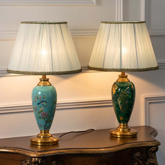 Turquoise ceramic table lamp with floral hand-painted design and pleated white shade turned on against white background creating warm soft lighting.