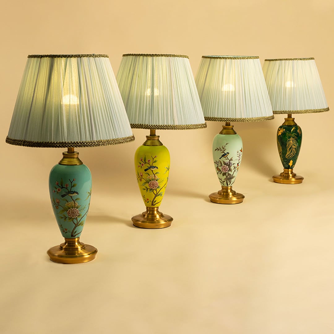 Collection of floral ceramic table lamps in mint green turquoise yellow and emerald green with gold bases and pleated white shades turned on together spreading warm elegant illumination.