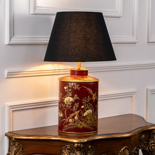 Red ceramic table lamp with floral bird artwork and black fabric shade placed on carved wooden console adding warm classic lighting.