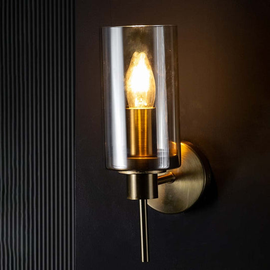 Contemporary wall sconce glowing warmly through a tall clear glass shade against a black background enhancing the warm light and polished metal finish.