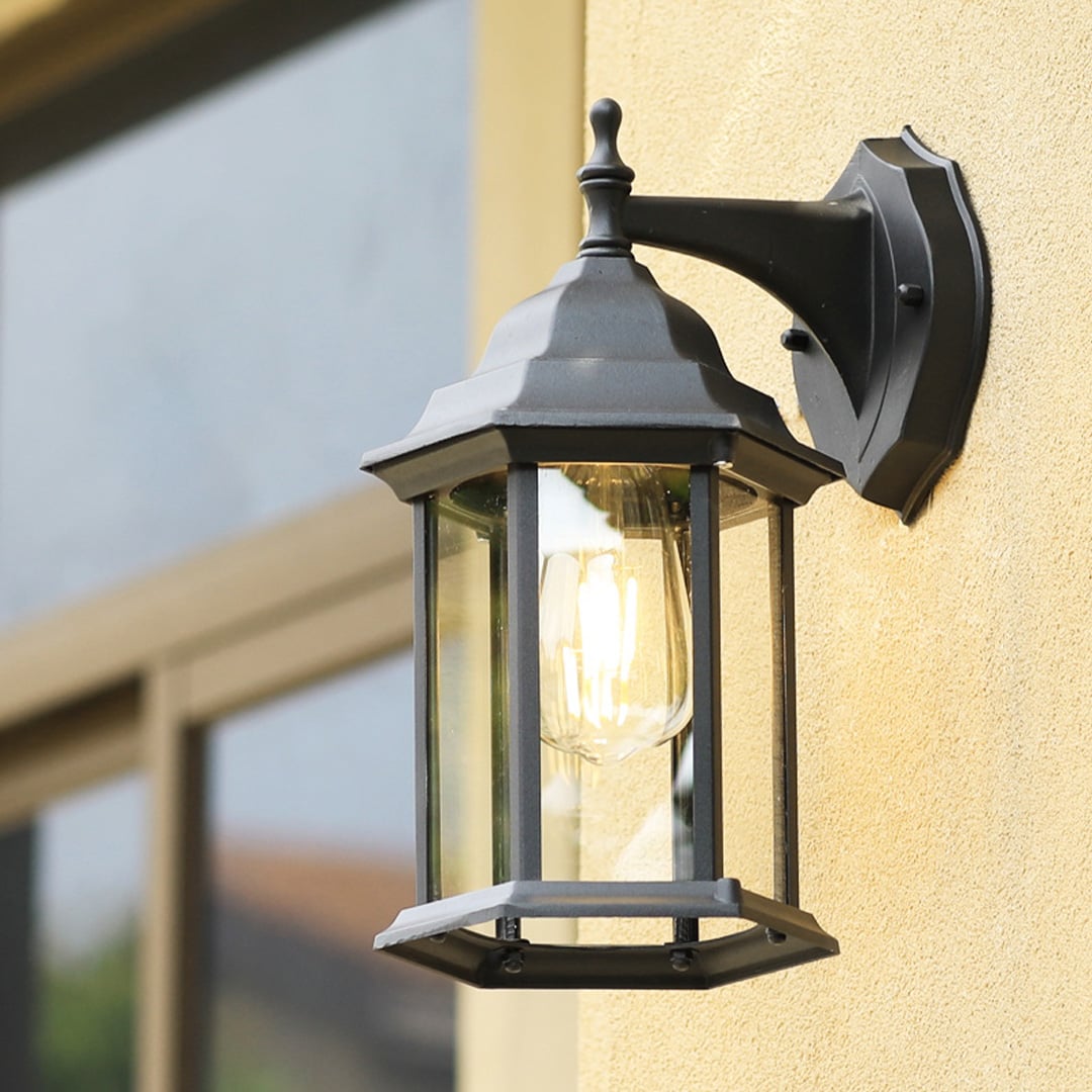 A decorative outdoor wall lantern with the light on mounted near a doorway casting a soft ambient glow on the wall and enhancing the natural greenery around the entrance.