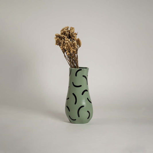 Tall green ceramic vase with black curved patterns holding dried flowers placed on a wooden chest to enhance a cozy vintage themed room.