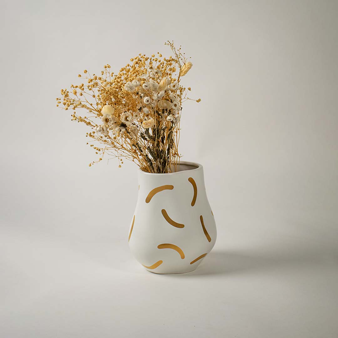 A softly shaped ceramic vase decorated with golden strokes shown in a minimal light filled setup adding a warm contemporary touch to the decor space.