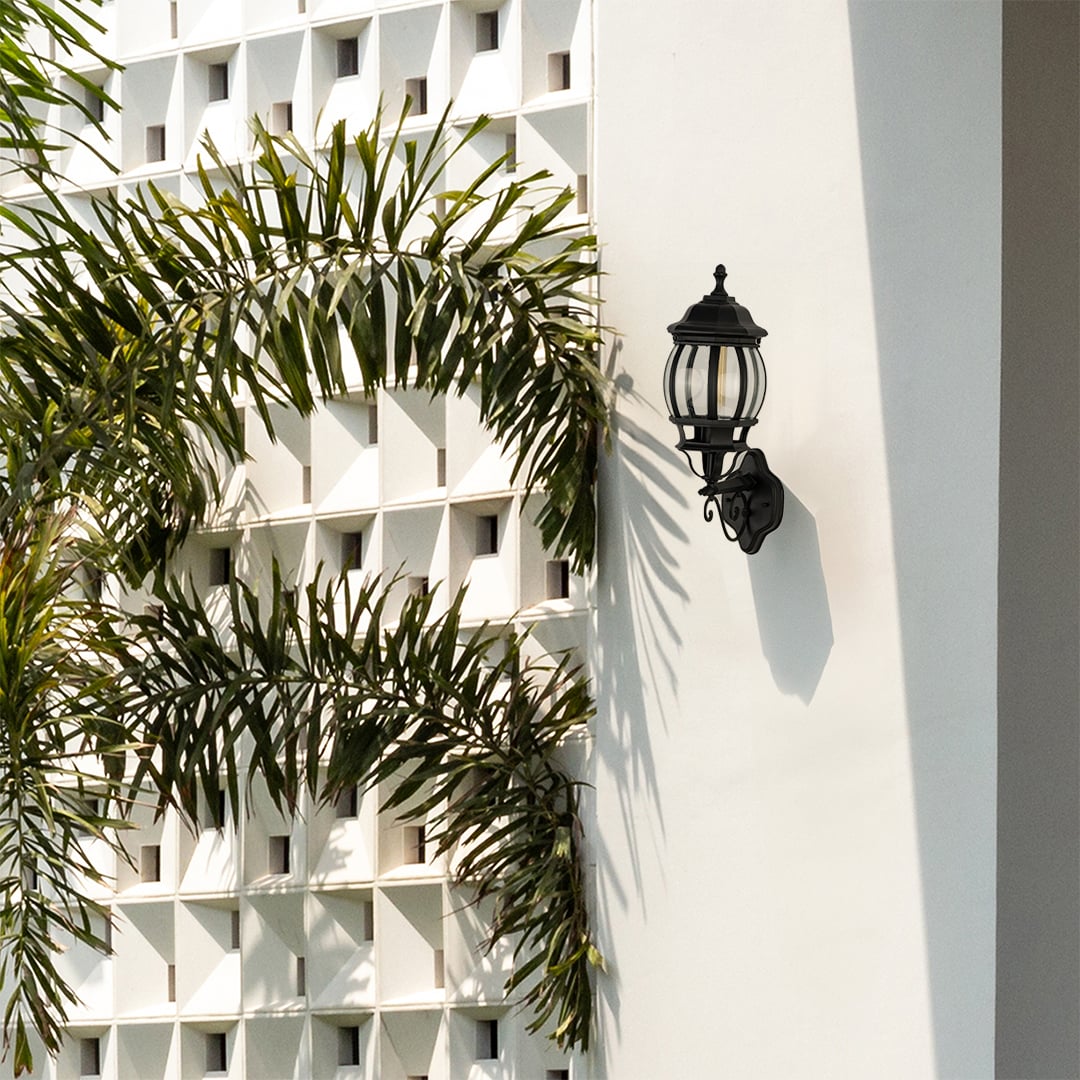 Black metal traditional wall lamp with vertical ribbed glass shade installed on white decorative breeze block wall near palm trees.