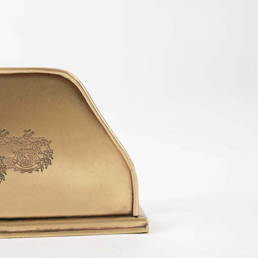 Detailed closeup view of the intricate engraved floral logo on the gold brass napkin holder surface highlighting craftsmanship.