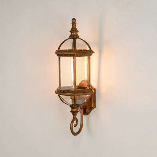 Antique brass metal outdoor wall light on white background with an elongated glass lantern and bottom scroll for decor.