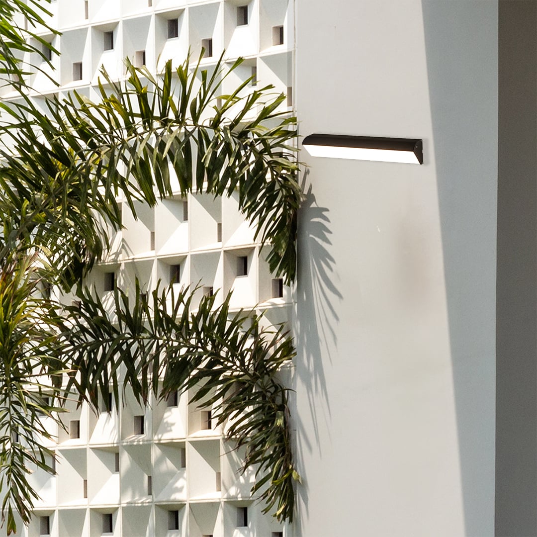 Sleek black horizontal LED wall lamp installed on white decorative breeze block wall near palm trees in a garden setting.