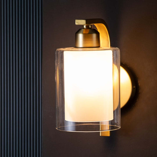 Elegant gold brass wall sconce featuring a dual clear and frosted glass cylinder shade fixture over a dark backdrop.