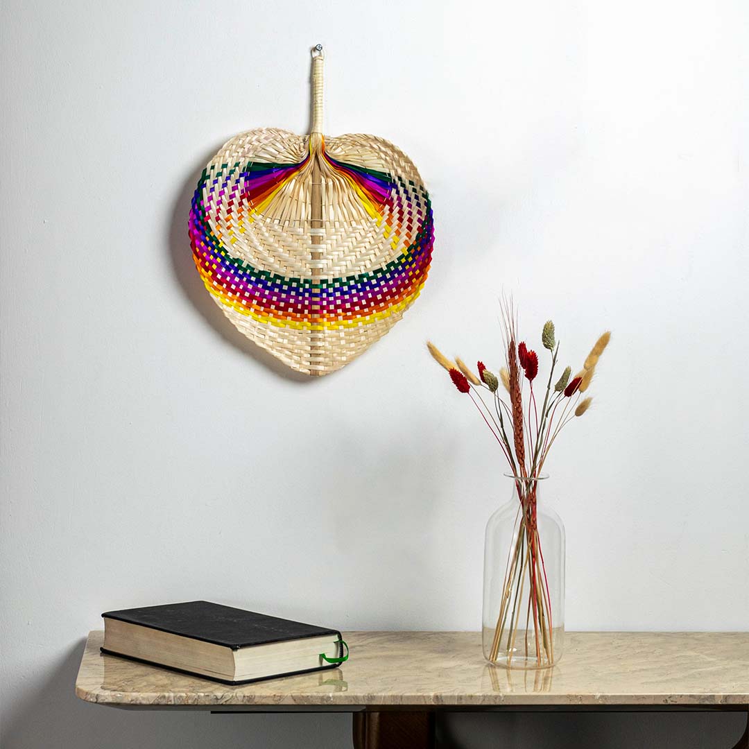 Rainbow bamboo hand fan hanging on wall creating colorful tropical bohemian accent in modern minimalist interior design space.