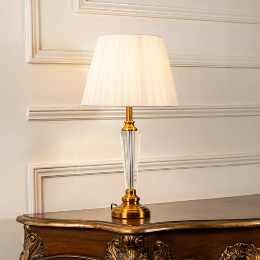 Crystal and gold table lamp with pleated shade turned on against white background perfect for bedside table lamp.