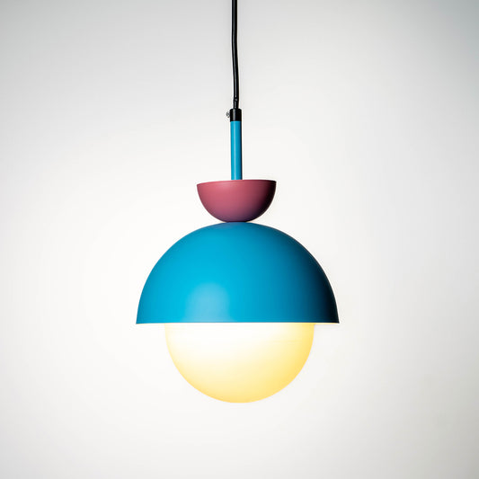Stylish dual tone pendant light in teal and pink metal finish with glowing glass globe shade hanging against white background for trendy decor spaces.