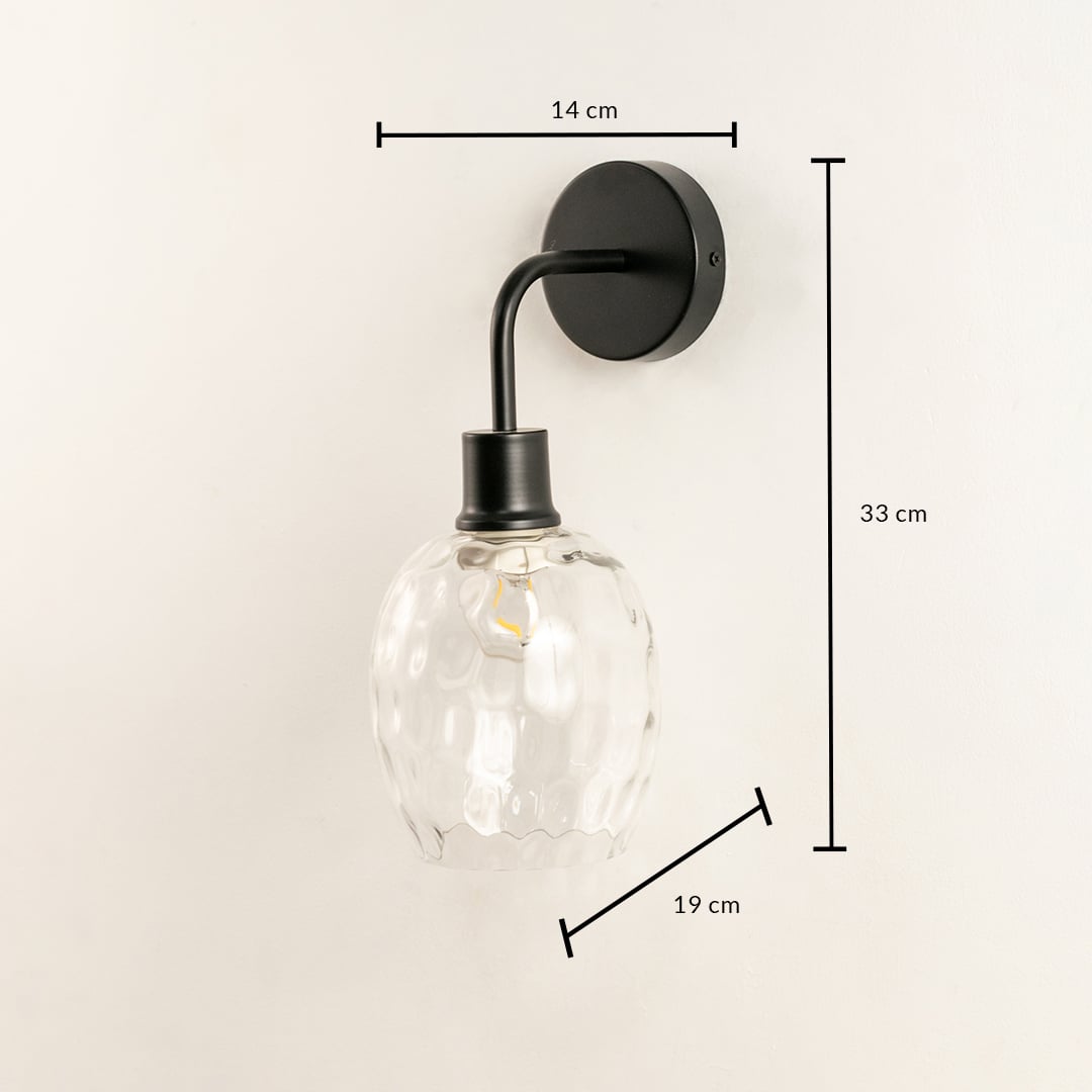 A glass and black metal wall light measuring 33 cm height 19 cm depth and 14 cm width glowing softly on a pale white background adding warm illumination and modern décor.