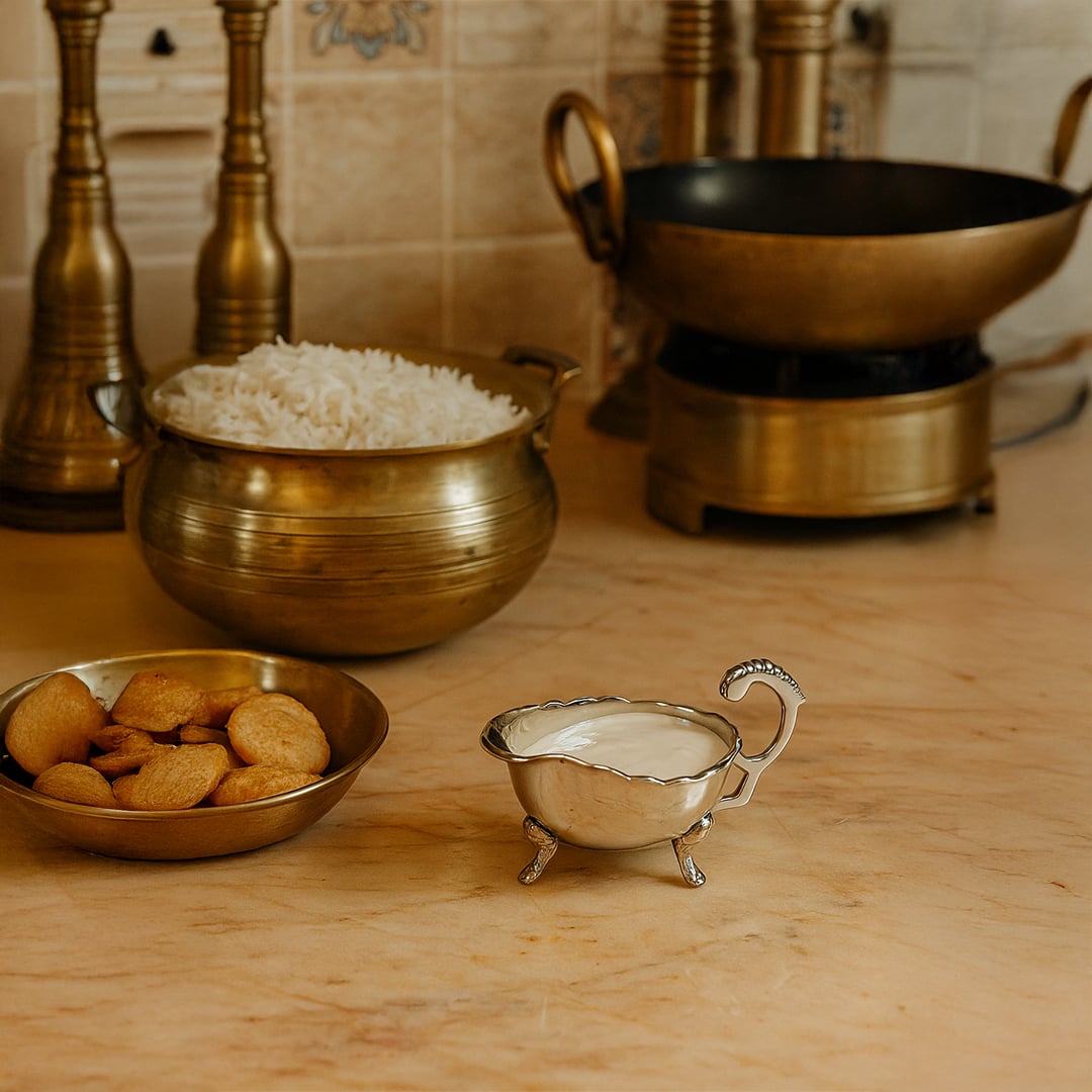 Elegant silver plated sauce bowl filled with white cream placed on a marble counter next to traditional brass cookware and rice handi.