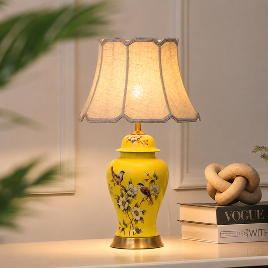 A yellow ceramic table lamp with a fluted shade, decorated with painted birds and flowers, shown lit on a tabletop next to books and a decorative knot.