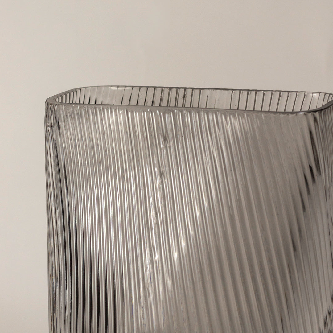 Tall clear ribbed glass vase closeup with vertical texture placed on wooden surface against neutral wall background enhancing modern decorative styling.