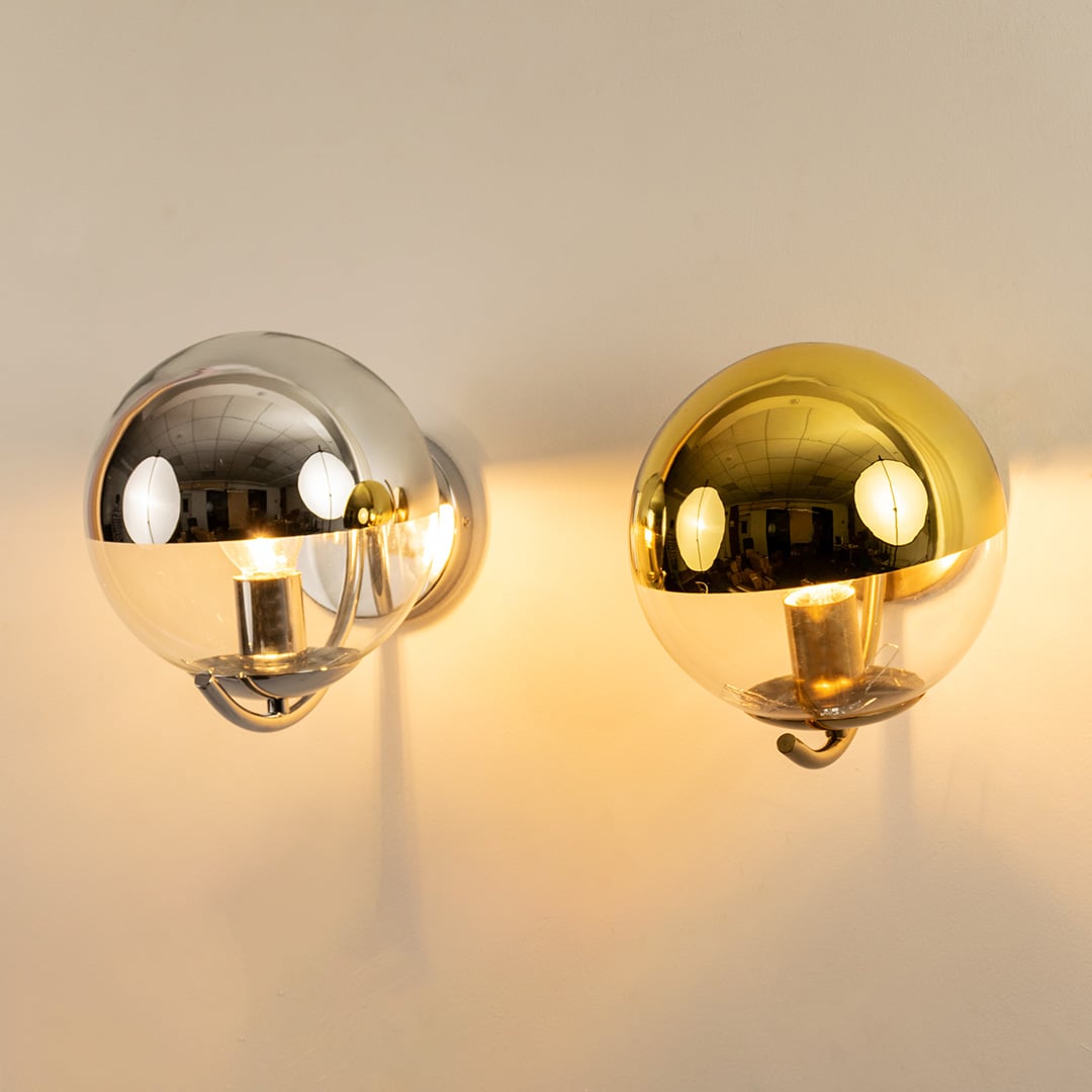 A duo of metal wall lights in chrome and gold finishes glowing warmly against a clean white wall creating reflective highlights and modern luxurious décor.