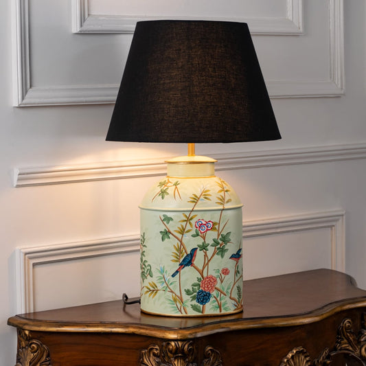 Table lamp with black fabric shade and cream ceramic base featuring colorful floral artwork placed on a wooden table against a white wall background.