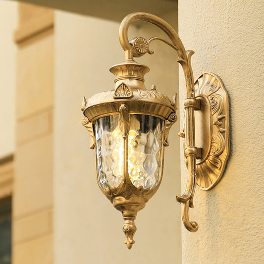 A gold antique wall lantern with the light on illuminating the exterior wall and casting a warm glow across the entrance creating an elegant and inviting outdoor ambiance.
