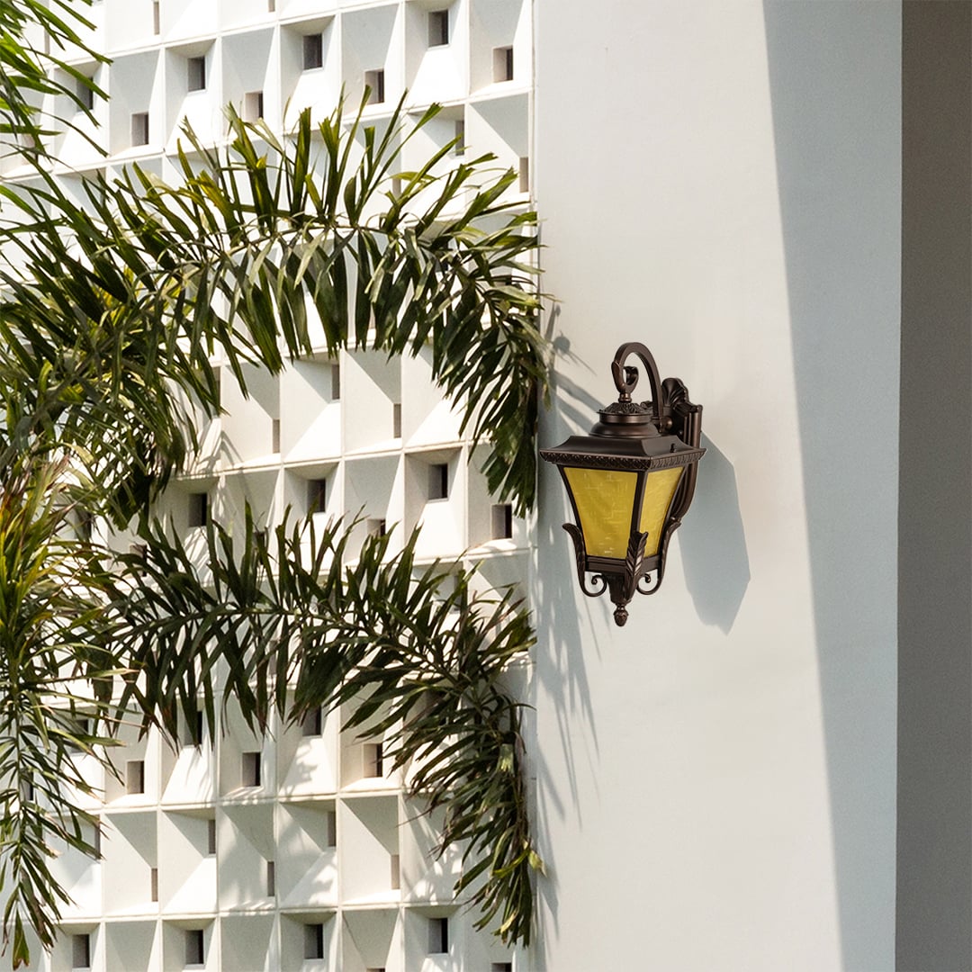 A bronze outdoor lantern with the light off mounted on a bright exterior wall surrounded by palm leaves highlighting its ornate frame frosted yellow glass and classic curved arm design.
