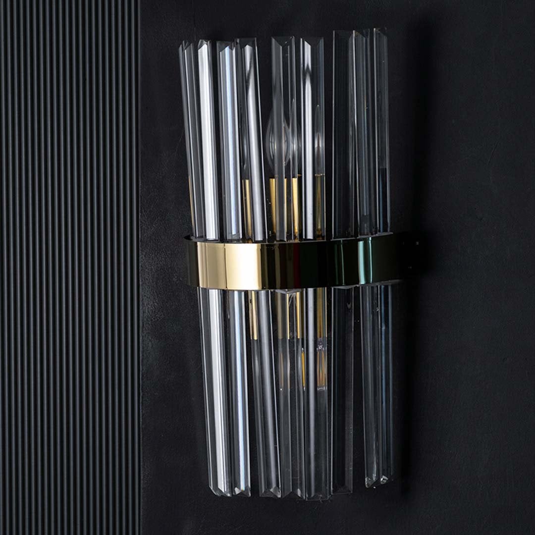 Crystal wall light with the light on against a black background creating a dramatic glow that enhances the reflective glass rods and golden band.