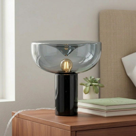 Stylish smoked glass table lamp with black metal base placed on wooden console against soft neutral wall background perfect for modern interior lighting.