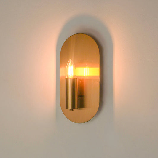 A modern wall sconce with an oval backplate and cylindrical metal fixture, illuminated with a warm yellow light against a plain off-white wall.
