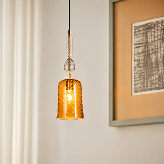 A hanging pendant light with a textured amber glass shade and a brass-toned chain, shown lit in a softly styled interior setting with a framed print on the wall.