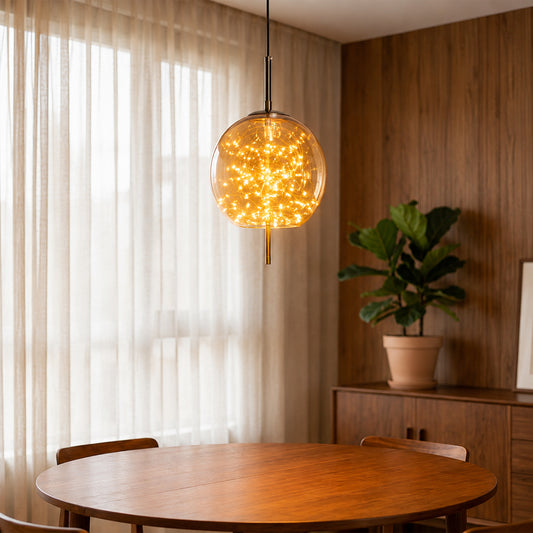A spherical glass pendant light filled with warm glowing fairy lights hangs over a round wooden dining table in a cozy room with sheer curtains, a wood-paneled wall, and a potted plant.