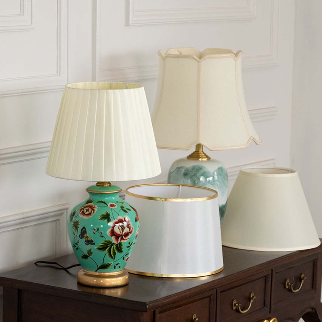 Green floral ceramic table lamp with pleated shade displayed on wooden console beside pastel lampshades