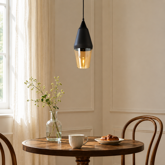 A modern black pendant light with a warm glowing glass shade hangs over a round wooden dining table in a softly lit room with cream walls, a window, sheer curtains, a glass vase with flowers, a white mug, and a croissant on a plate.