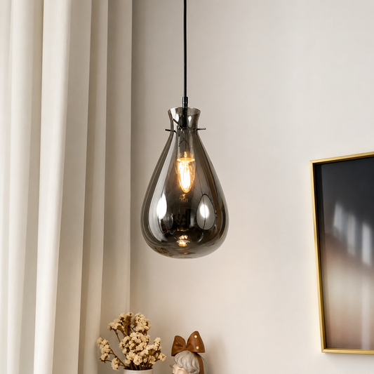 A teardrop-shaped smoked glass pendant light hanging in a modern room setting with a black cord, warm glowing bulb, cream curtains, and a framed print on the wall.
