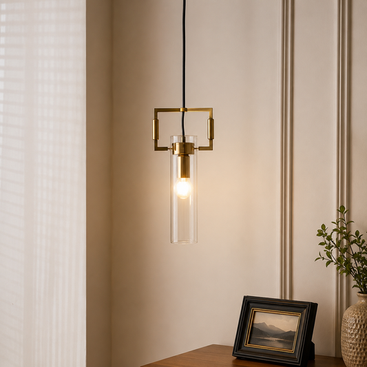 A modern brass pendant light with a clear glass cylinder shade hanging in a warm, lit interior setting next to a window and a small framed picture.