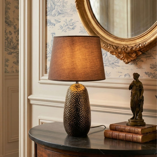 A table lamp with a textured dark brown and gold patterned base and a warm brown fabric drum shade, styled on a dark wood console table in an elegant room with wallpaper, a gold mirror, and a small bronze statue.