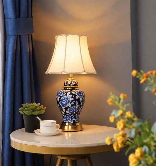 A decorative table lamp with a blue and white floral ceramic base and a cream pleated shade, styled on a round marble-top side table in a room with blue curtains and flowers.